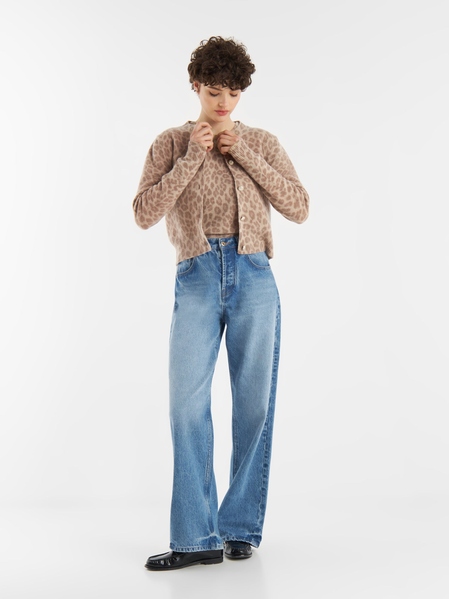 Eine Person mit kurzen lockigen Haaren trägt die hellbraune Strickjacke Leo Print aus weicher Merinowolle, gepaart mit hochhackigen Bluejeans und schwarzen Schuhen. Sie steht vor einem weißen Hintergrund und schaut nach unten, während sie die Strickjacke zuknöpft.
