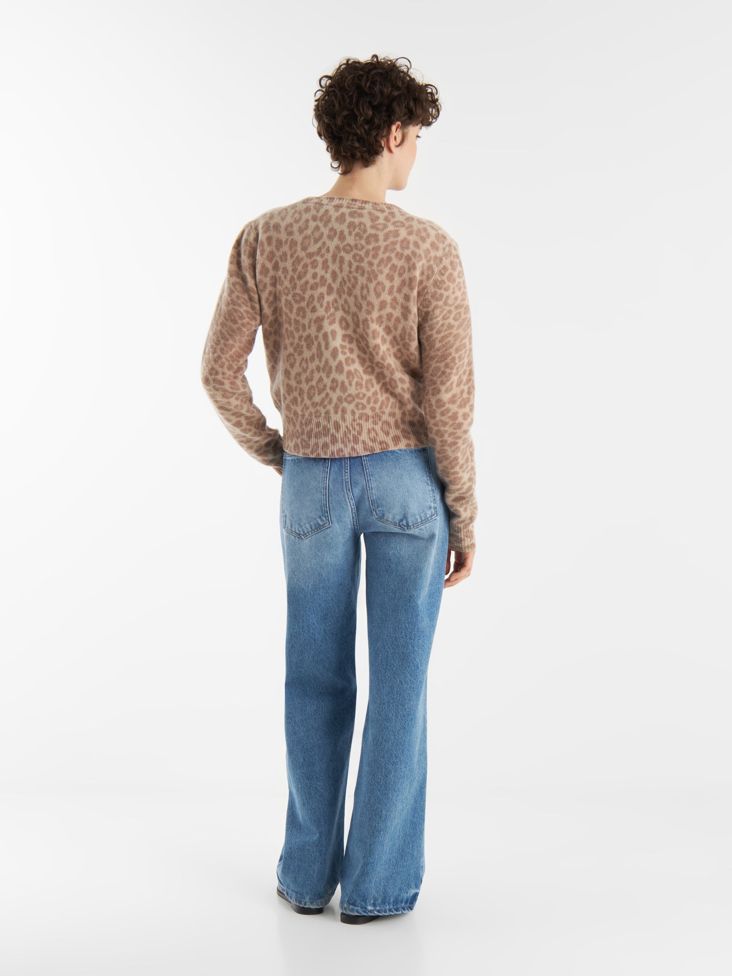 Eine Person mit kurzen lockigen Haaren, die den Cardigan Leo Print aus kuscheligen Naturfasern und hellblaue Schlagjeans trägt, steht mit dem Gesicht zur Wand vor einem schlichten weißen Hintergrund.