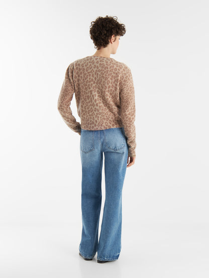 Eine Person mit kurzen lockigen Haaren, die den Cardigan Leo Print aus kuscheligen Naturfasern und hellblaue Schlagjeans trägt, steht mit dem Gesicht zur Wand vor einem schlichten weißen Hintergrund.