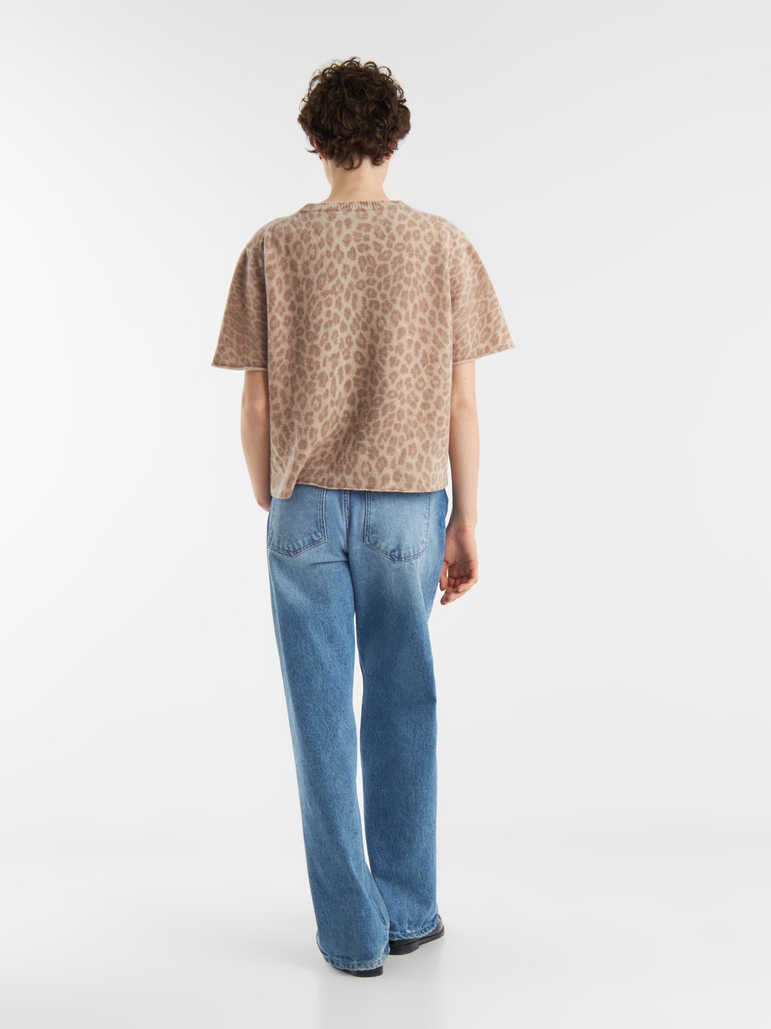 Eine Person mit kurzen lockigen Haaren, die das beigefarbene T-Shirt von Leo Strick aus Italien und eine blaue Jeans trägt, steht mit dem Gesicht nach hinten auf einem schlichten weißen Hintergrund.
