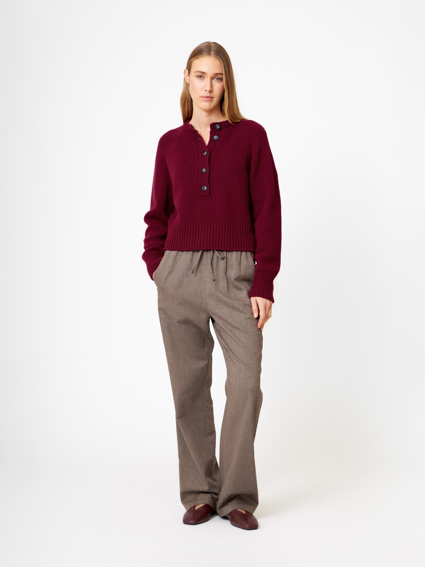 Eine Frau mit glattem, hellbraunem Haar trägt den Pullover Raglan Burgundy, eine braune Hose mit weitem Bein und burgunderfarbene Schuhe vor einem schlichten weißen Hintergrund. Sie hat einen neutralen Gesichtsausdruck.