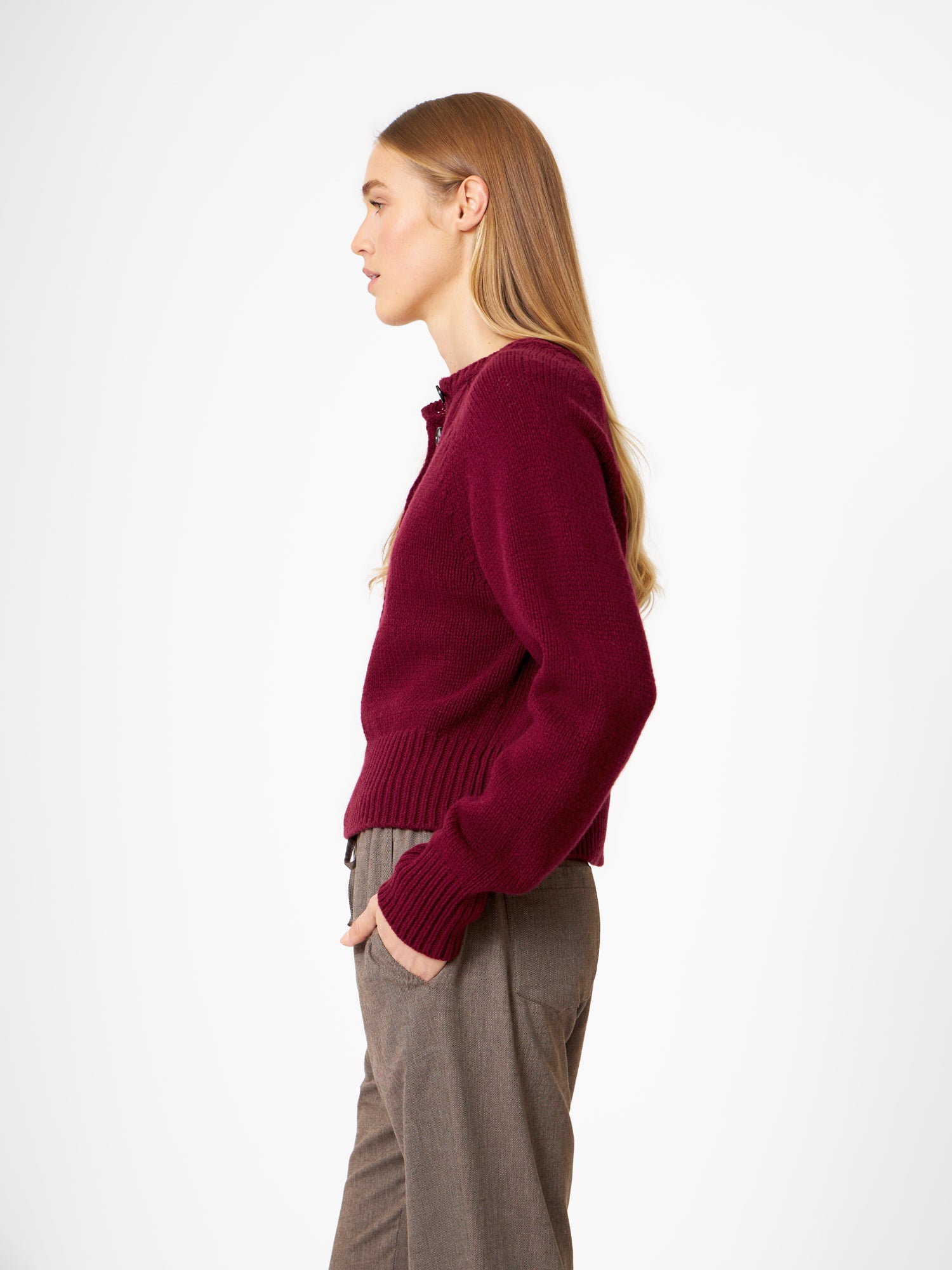 Eine Frau mit langem, glattem blondem Haar steht im Profil und trägt einen Pullover Raglan Burgundy und eine braune Hose vor einem schlichten weißen Hintergrund.
