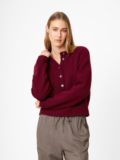 Eine Frau mit glattem blondem Haar trägt einen Pullover Raglan Burgundy und eine graue Hose mit Kordelzug. Sie steht vor einem schlichten weißen Hintergrund und hat einen neutralen Ausdruck.