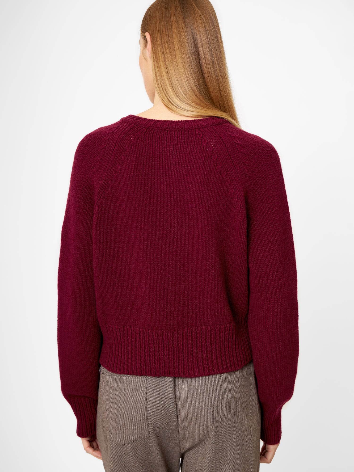 Eine Person mit langem, glattem, hellbraunem Haar wird von hinten gezeigt. Sie trägt einen burgunderroten Pullover und eine braune Hose und steht vor einem schlichten weißen Hintergrund.