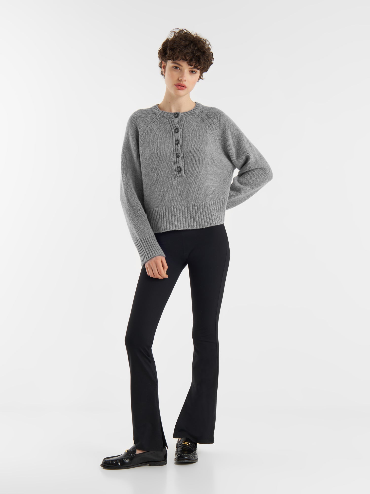 Eine Person mit kurzen lockigen Haaren und einer entspannten Pose steht vor einem weißen Hintergrund und trägt den grauen Pullover Raglan, eine schwarze Schlaghose und schwarze Slipper.