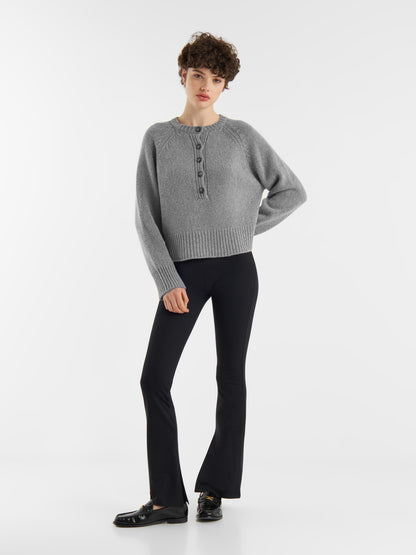 Eine Person mit kurzen lockigen Haaren und einer entspannten Pose steht vor einem weißen Hintergrund und trägt den grauen Pullover Raglan, eine schwarze Schlaghose und schwarze Slipper.