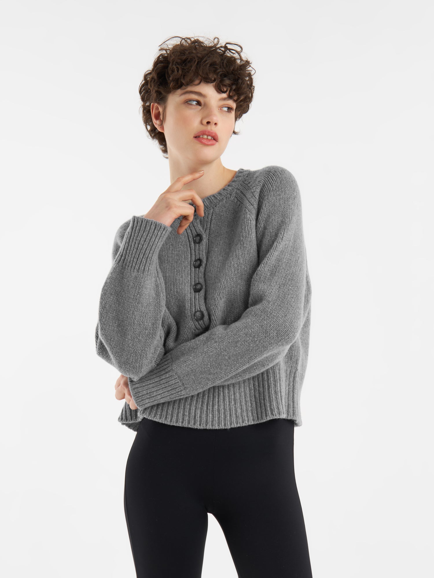 Eine Person, die einen grauen Pullover Raglan und schwarze Leggings trägt, steht vor einem weißen Hintergrund und schaut zur Seite, wobei eine Hand ihr Kinn berührt.