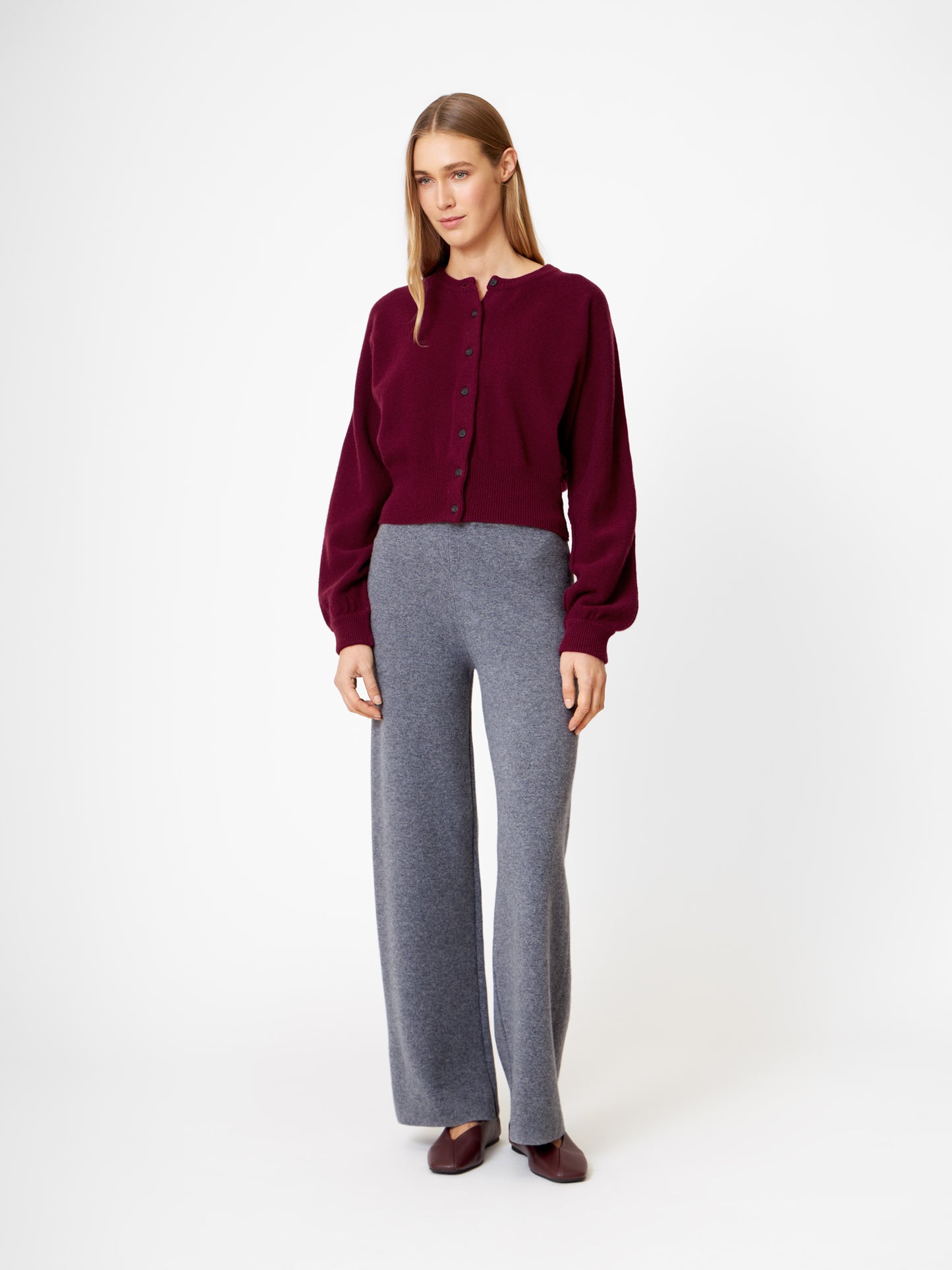 Eine Frau mit glattem, schulterlangem Haar und neutralem Gesichtsausdruck steht vor einem weißen Hintergrund und trägt die Strickjacke Charlotte Burgundy, eine graue Hose mit weitem Bein und burgunderfarbene Schuhe.