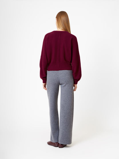 Eine Person mit glattem, langem Haar steht mit dem Gesicht nach hinten und trägt die Strickjacke Charlotte Burgundy, eine graue Hose mit weitem Bein und braune Schuhe vor einem schlichten weißen Hintergrund.