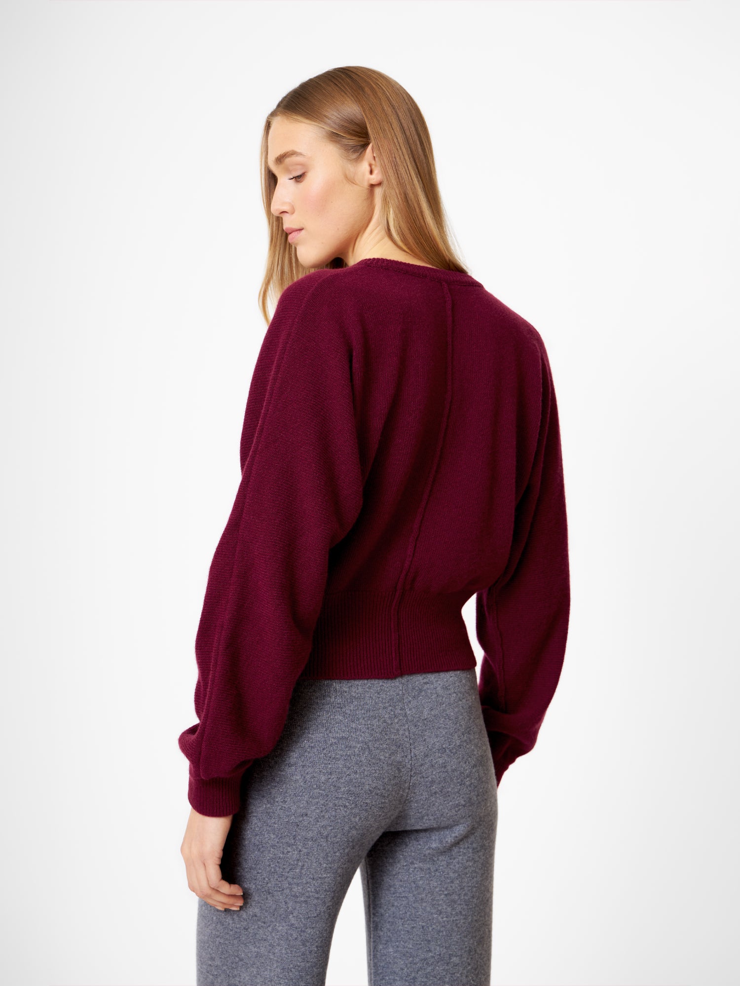 Eine Frau mit langem, glattem blondem Haar trägt die Strickjacke Charlotte Burgundy und eine graue Hose mit hoher Taille. Sie steht und schaut über ihre Schulter vor einem schlichten weißen Hintergrund.