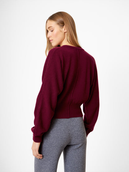 Eine Frau mit langem, glattem blondem Haar trägt die Strickjacke Charlotte Burgundy und eine graue Hose mit hoher Taille. Sie steht und schaut über ihre Schulter vor einem schlichten weißen Hintergrund.