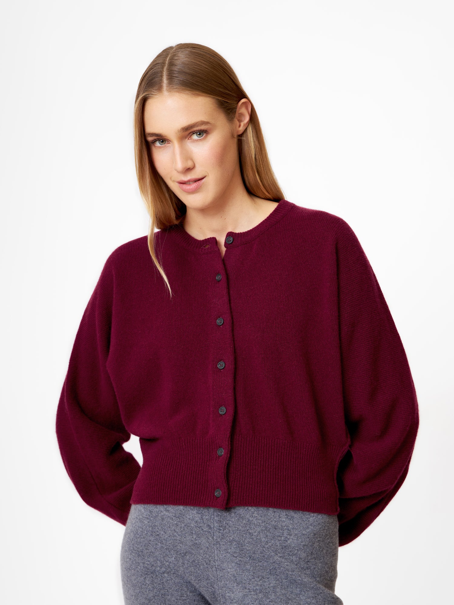 Eine Frau mit glattem, hellbraunem Haar trägt die locker sitzende Strickjacke Charlotte Burgundy und eine graue Hose, steht vor einem schlichten weißen Hintergrund und lächelt leicht in die Kamera.