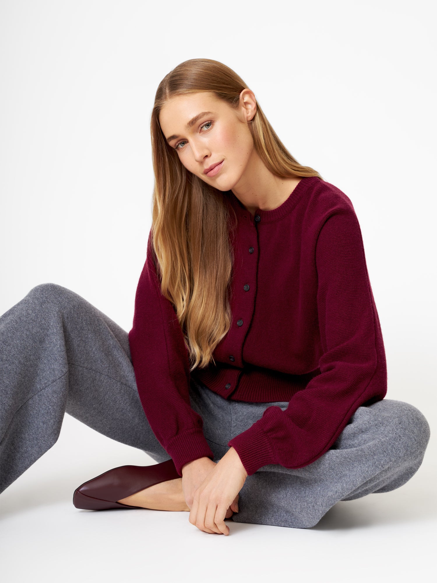 Eine Frau mit langem, glattem hellbraunem Haar sitzt im Schneidersitz auf dem Boden. Sie trägt die Strickjacke Charlotte Burgundy, eine graue Hose mit weitem Bein und burgunderfarbene Schuhe vor einem schlichten weißen Hintergrund.