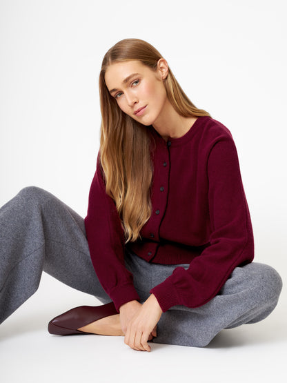 Eine Frau mit langem, glattem hellbraunem Haar sitzt im Schneidersitz auf dem Boden. Sie trägt die Strickjacke Charlotte Burgundy, eine graue Hose mit weitem Bein und burgunderfarbene Schuhe vor einem schlichten weißen Hintergrund.