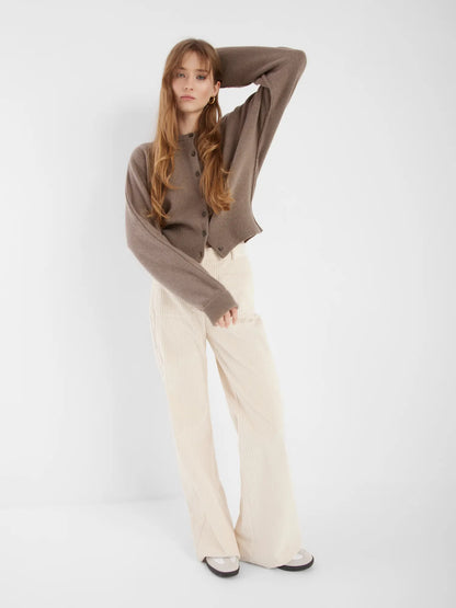 Eine Frau mit langen braunen Haaren steht vor einem weißen Hintergrund und trägt die Strickjacke Charlotte Camel, eine cremefarbene Hose mit weitem Bein und weiße Schuhe. Sie posiert mit einem erhobenen Arm und einem entspannten Gesichtsausdruck.