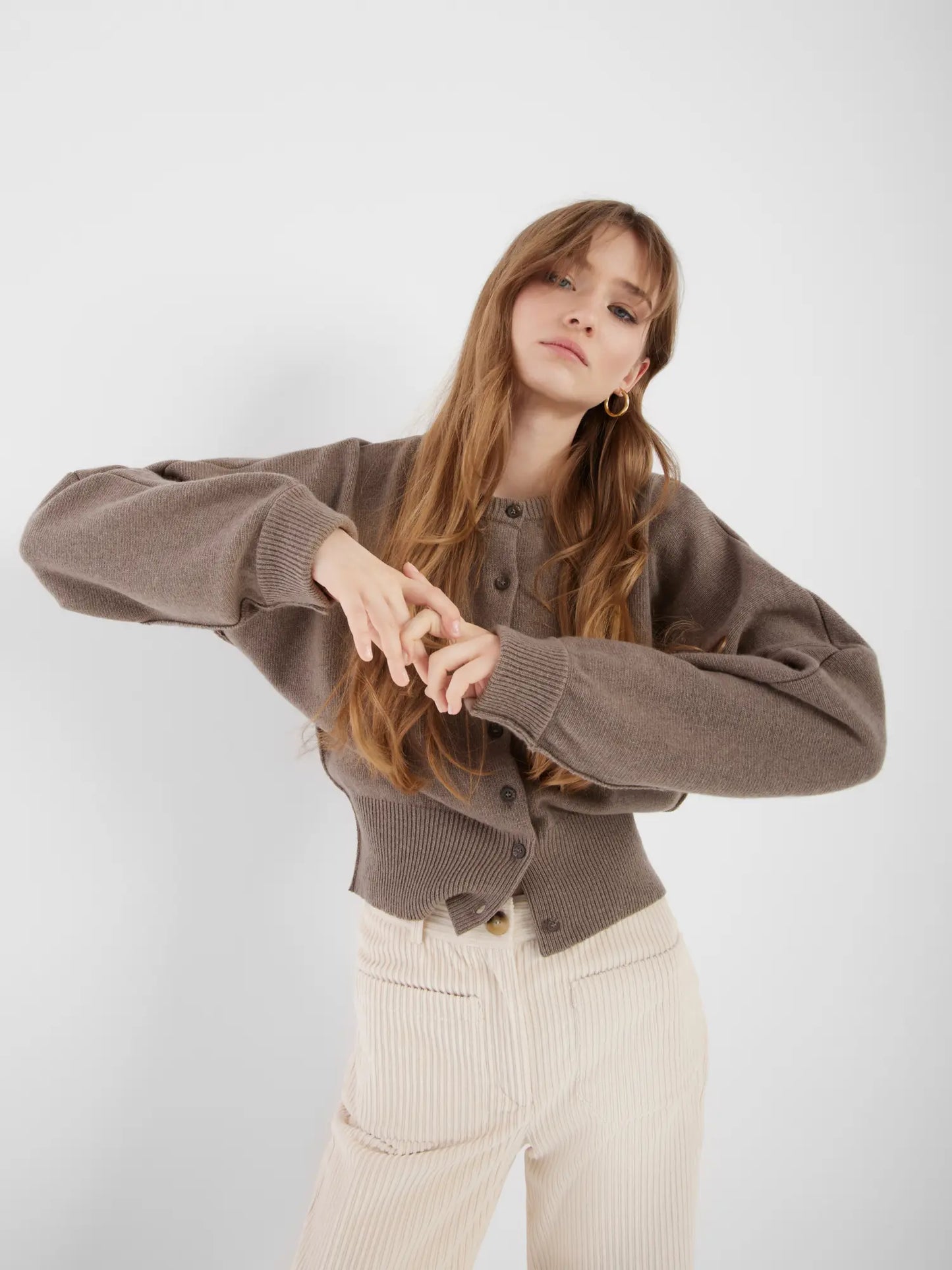 Eine junge Frau mit langem, hellbraunem Haar posiert vor einem schlichten weißen Hintergrund. Sie trägt die Strickjacke Charlotte Camel und eine cremefarbene Cordhose mit hoher Taille. Ihre Arme sind angewinkelt und die Hände vor sich verschränkt.