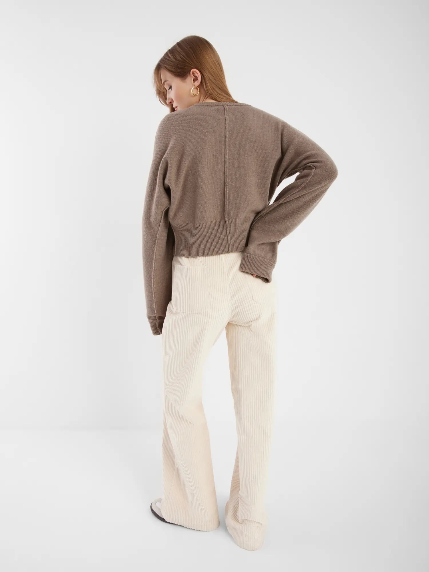 Eine Frau steht in einer Strickjacke Charlotte Camel und einer cremefarbenen Cordhose vor einem schlichten weißen Hintergrund.