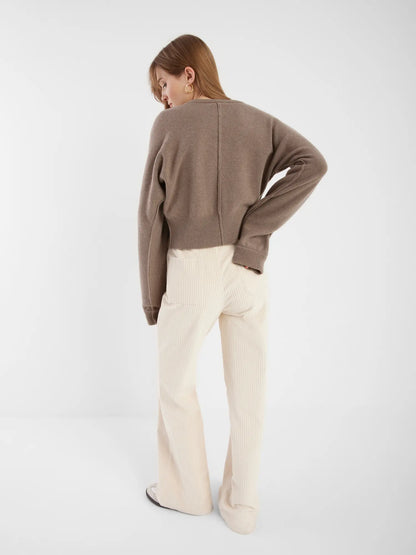 Eine Frau steht in einer Strickjacke Charlotte Camel und einer cremefarbenen Cordhose vor einem schlichten weißen Hintergrund.