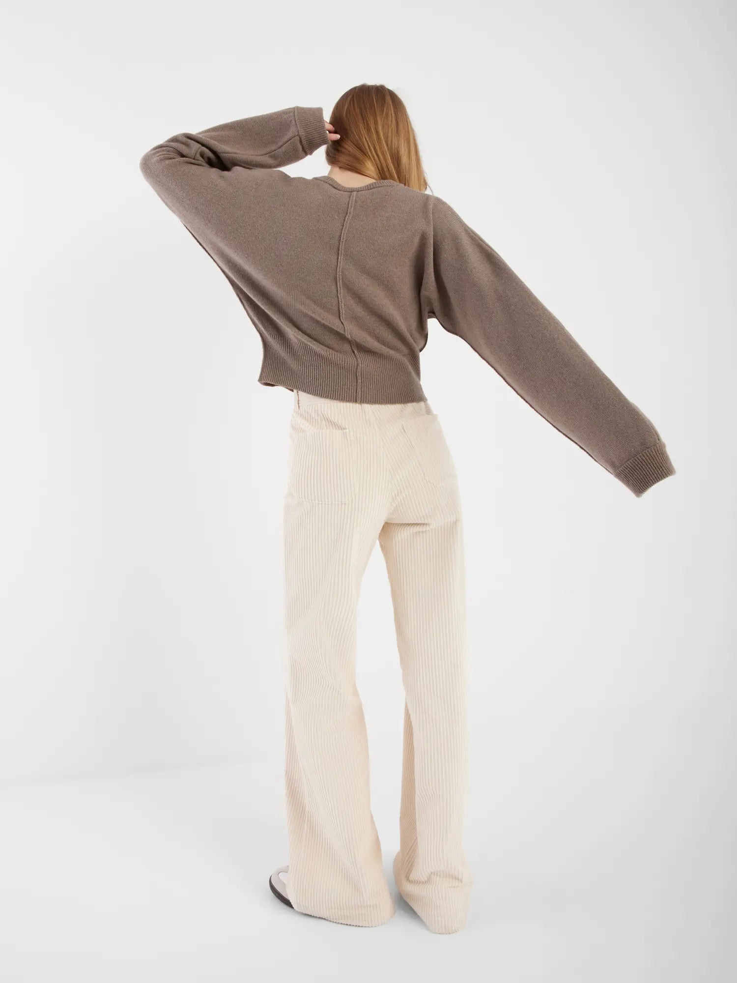 Eine Person mit langen Haaren steht mit dem Gesicht nach hinten, trägt die Strickjacke Charlotte Camel und eine cremefarbene Hose mit weitem Bein, einen Arm erhoben und den anderen an der Seite, vor einem schlichten weißen Hintergrund.