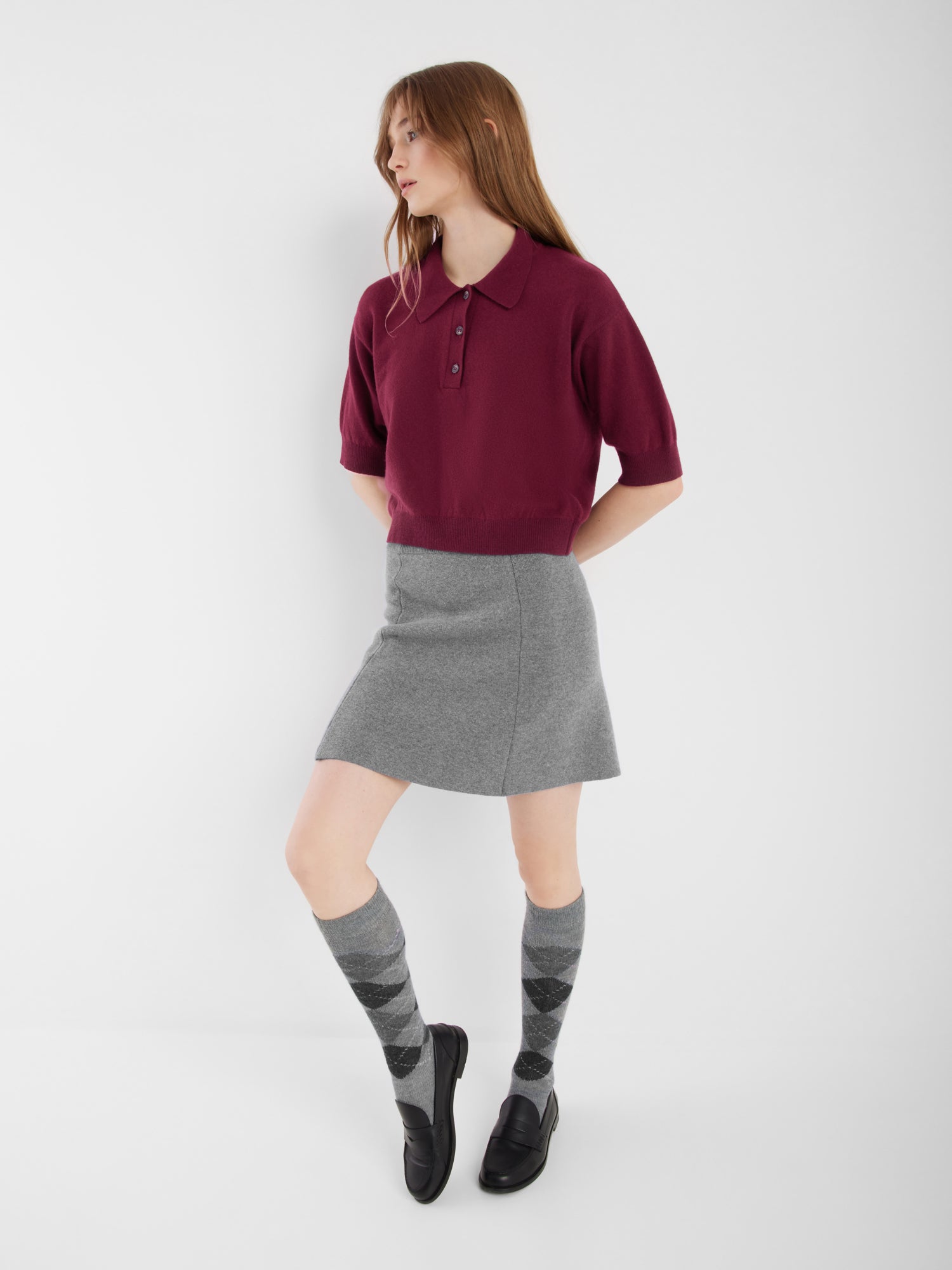 Eine Person mit langem glattem Haar trägt ein kurzärmeliges kastanienbraunes Polo, den Rock Marino Mini Grey, kniehohe Argyle-Socken und schwarze Slipper, während sie vor einem schlichten weißen Hintergrund steht.