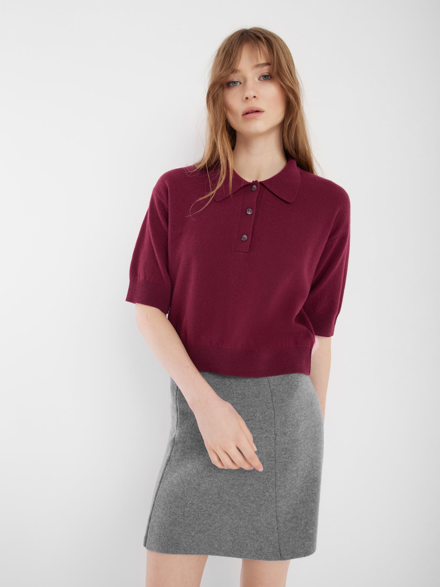 Eine junge Frau mit langen hellbraunen Haaren trägt ein kurzärmeliges bordeauxfarbenes Poloshirt und einen Rock Marino Mini Grey vor einem schlichten weißen Hintergrund.