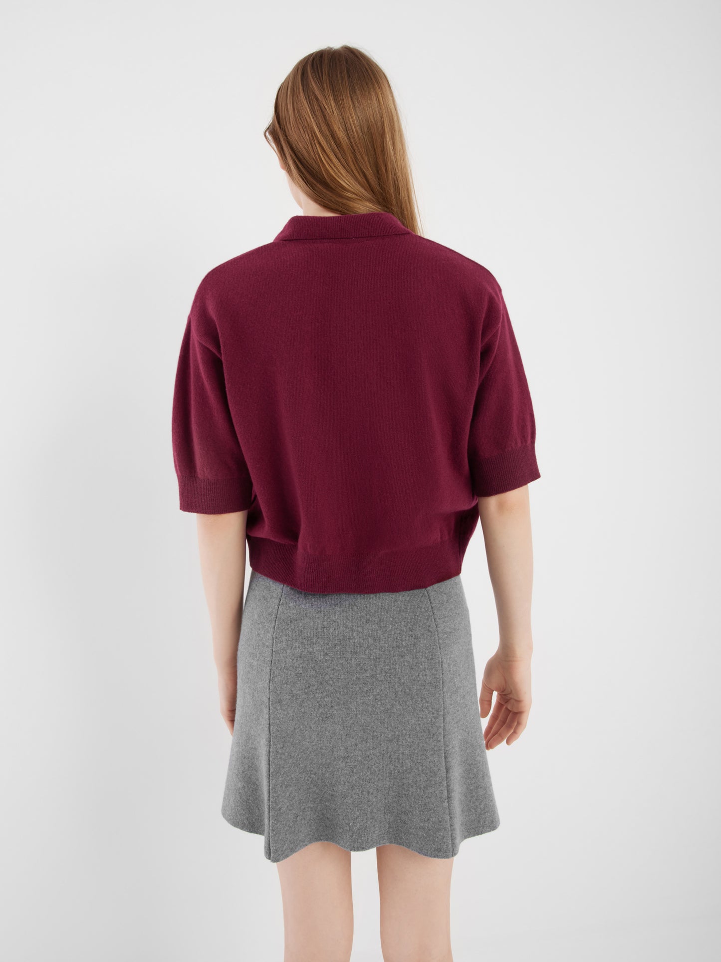 Eine Frau mit langem, hellbraunem Haar ist von hinten zu sehen. Sie trägt einen weinroten Pullover und einen grauen ausgestellten Rock vor einem schlichten weißen Hintergrund.