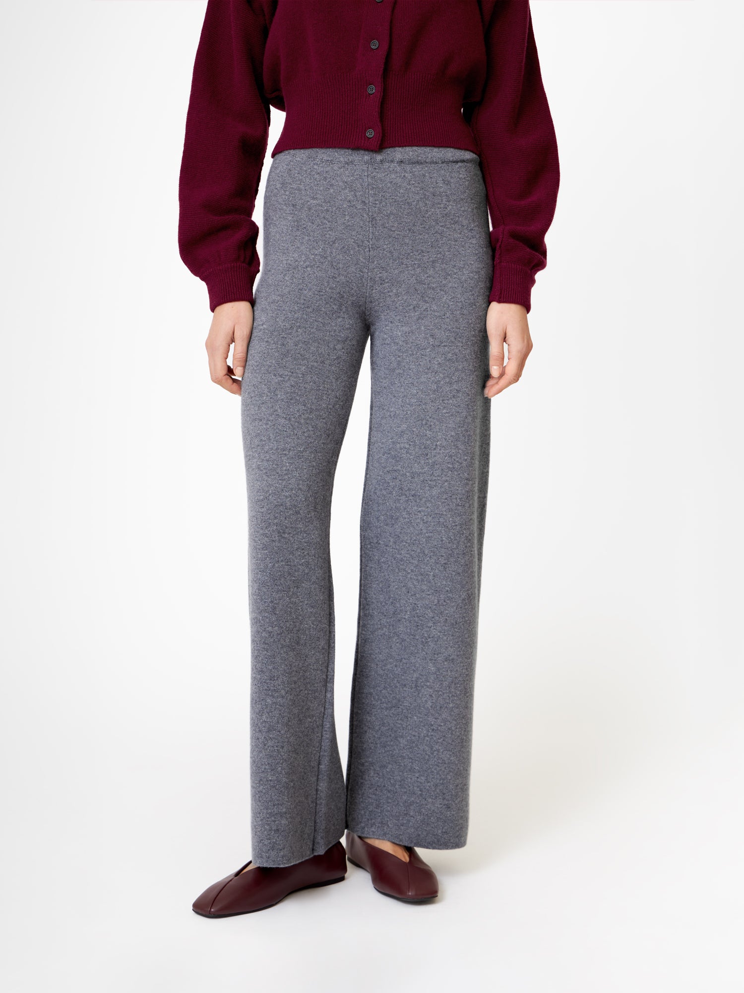 Eine Person trägt eine bordeauxfarbene Strickjacke mit Knöpfen, eine graue Hose aus Merino Grey-Strick - ideal als Wollhose Damen - und dazu passende bordeauxfarbene Slipper, die vor einem schlichten weißen Hintergrund stehen. Der Kopf ist auf dem Bild nicht zu sehen.