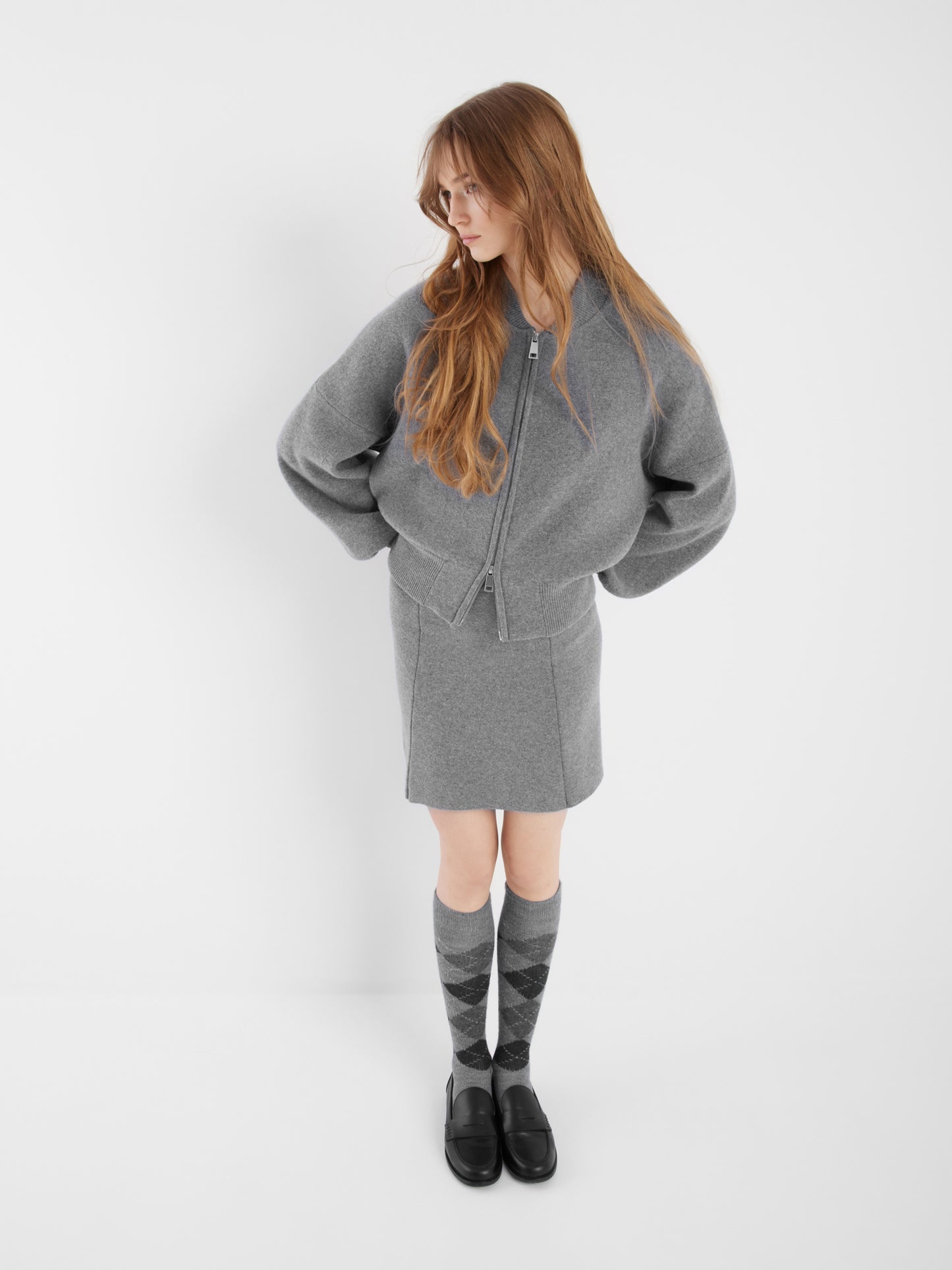 Eine Person mit langem, hellbraunem Haar trägt eine graue Bomberjacke, einen passenden Rock, Kniestrümpfe mit Argylemuster und schwarze Schuhe. Sie steht vor einem schlichten weißen Hintergrund und blickt zur Seite.