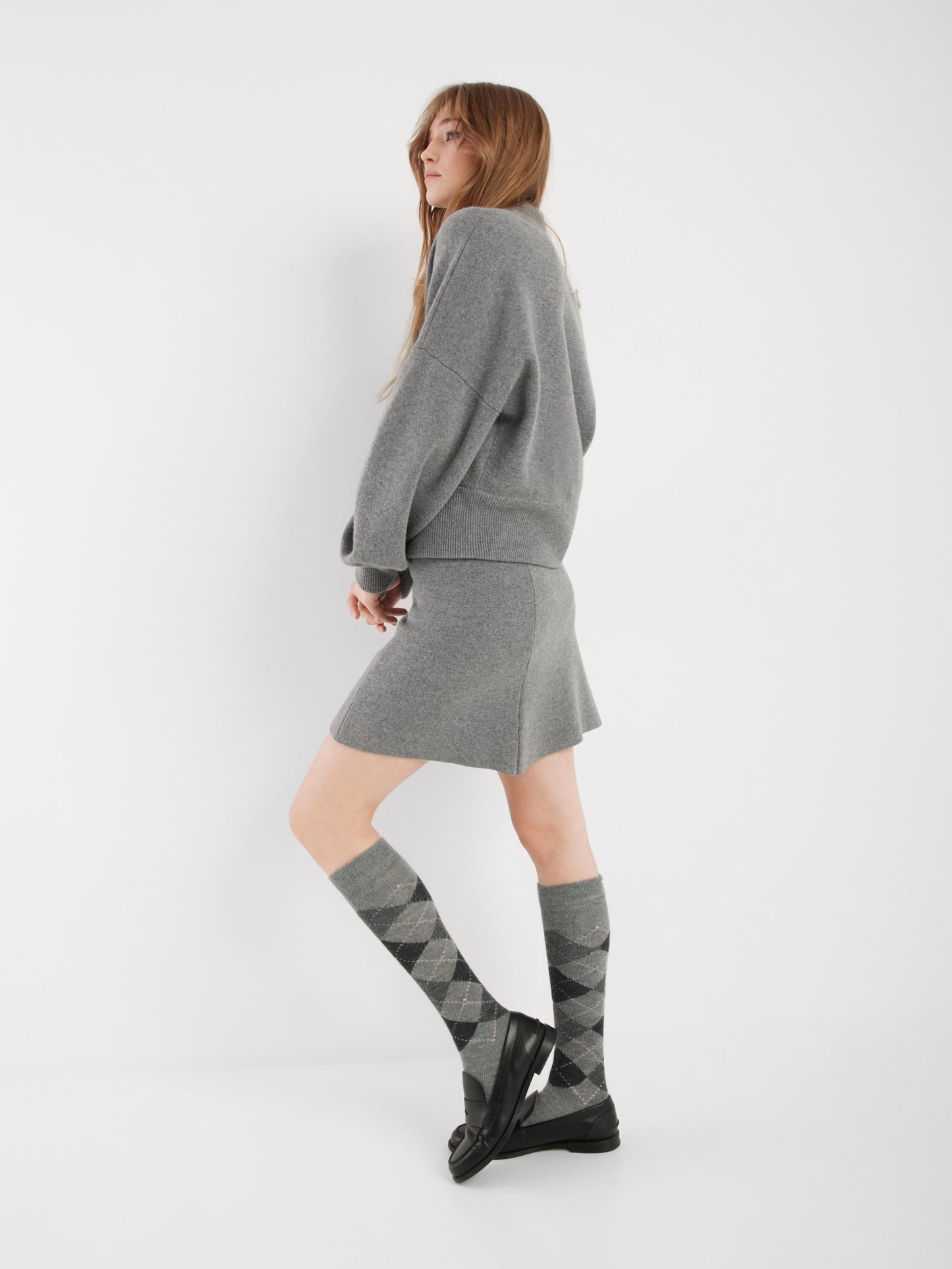 Eine Frau mit langen Haaren steht seitlich vor einem weißen Hintergrund. Sie trägt eine lockere graue Bomberjacke und einen Rock, Kniestrümpfe in Argyle-Optik und schwarze Slipper.