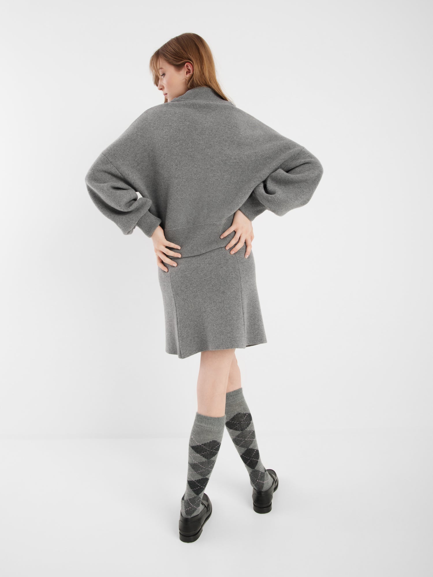 Eine Frau steht mit dem Rücken zur Kamera und trägt eine graue Bomberjacke, einen dazu passenden Rock, Kniestrümpfe in Argyle-Optik und schwarze Schuhe vor einem schlichten weißen Hintergrund.