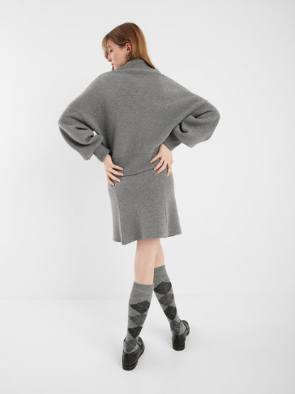 Eine Frau steht mit dem Rücken zur Kamera und trägt eine graue Bomberjacke, einen dazu passenden Rock, Kniestrümpfe in Argyle-Optik und schwarze Schuhe vor einem schlichten weißen Hintergrund.