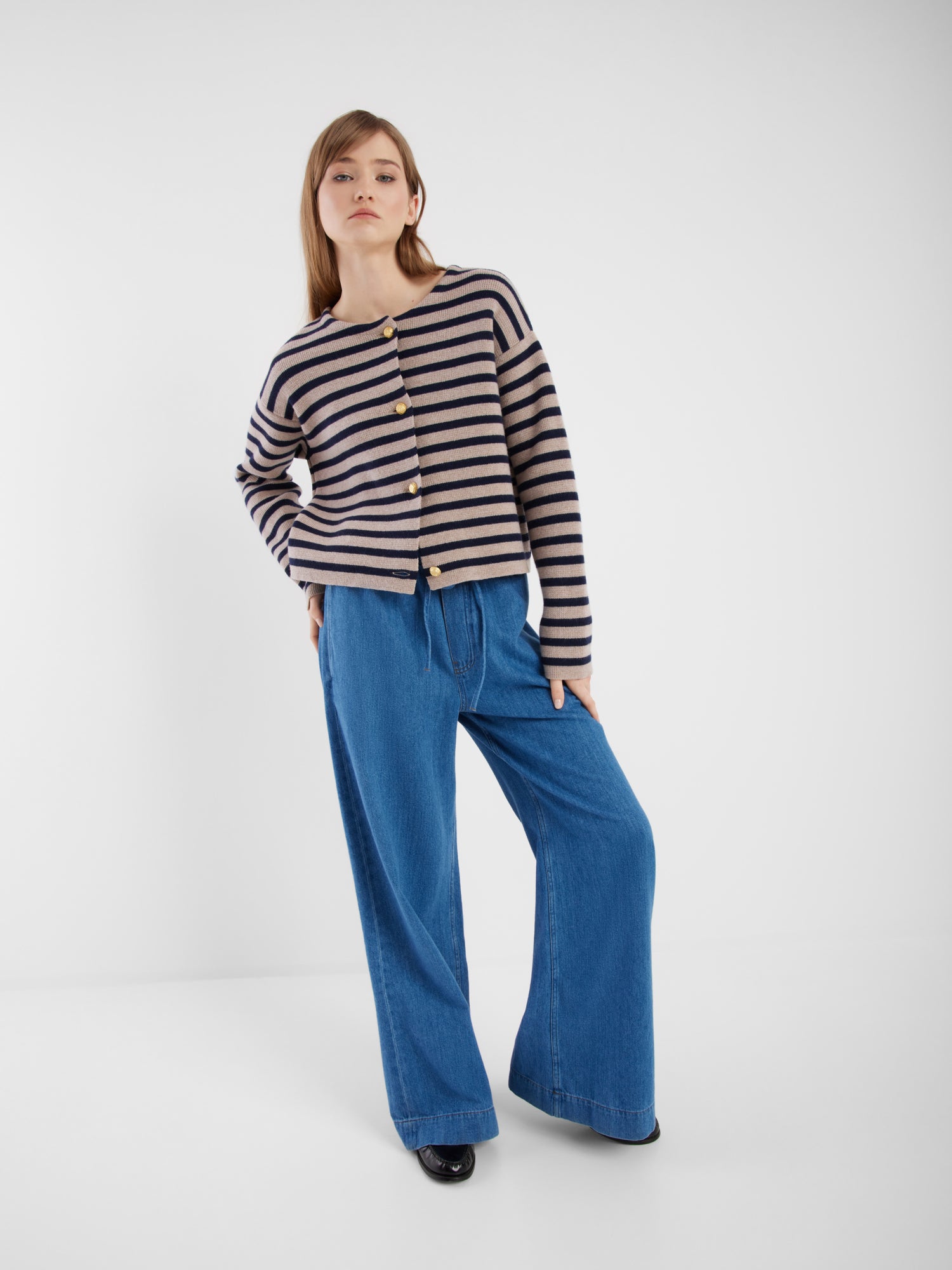 Eine Frau steht in einem weißen Raum und trägt die blau-beige gestreifte Jacke Sailor mit Knöpfen und weit geschnittenen Blue Jeans. Sie hat glattes, schulterlanges Haar und posiert entspannt mit einer Hand in der Tasche.