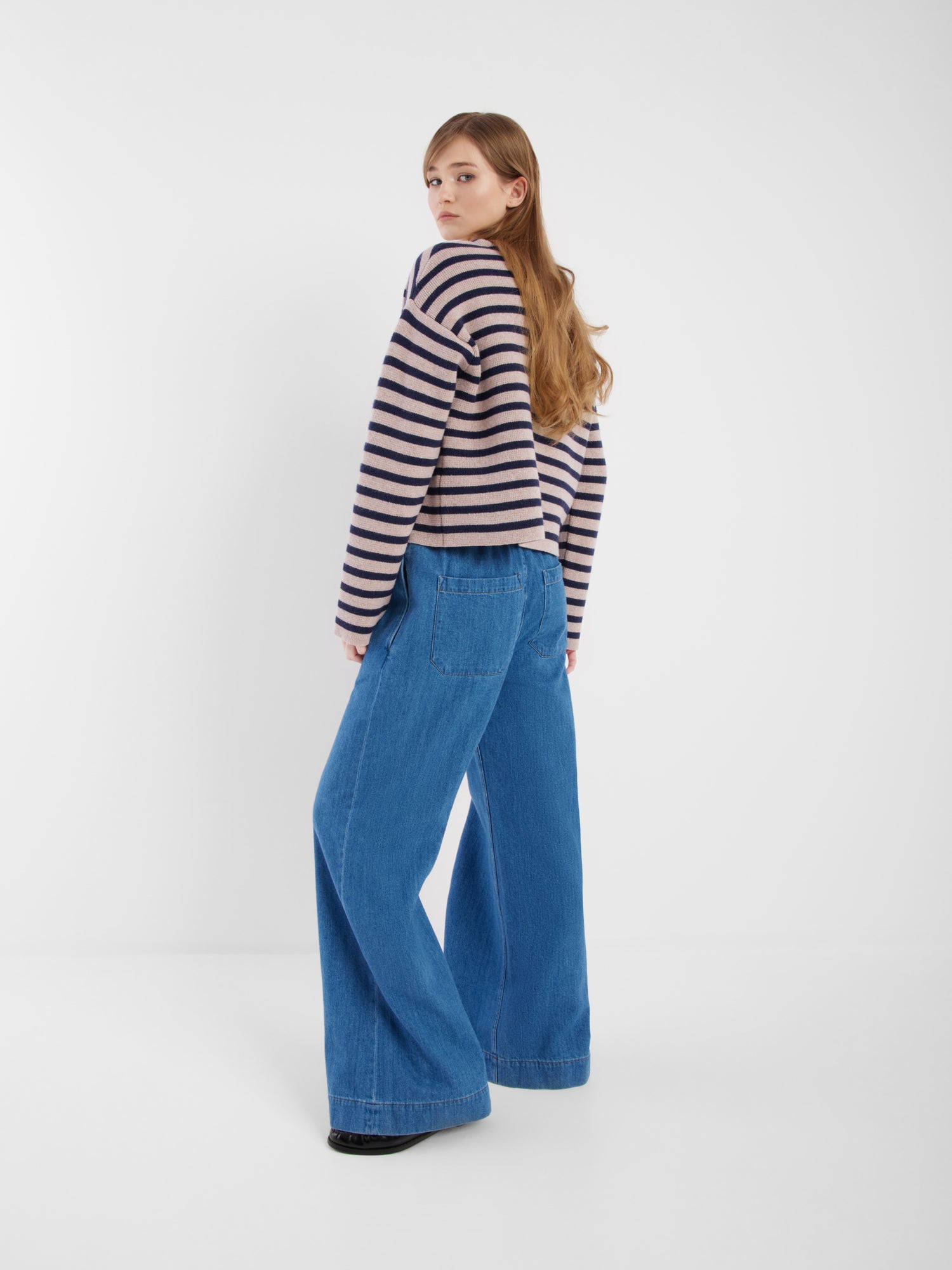 Eine Frau mit langen Haaren trägt die blau-beige gestreifte Jacke Sailor und eine weit geschnittene Bluejeans. Sie steht in einem minimalen weißen Raum und schaut über die Schulter in die Kamera.