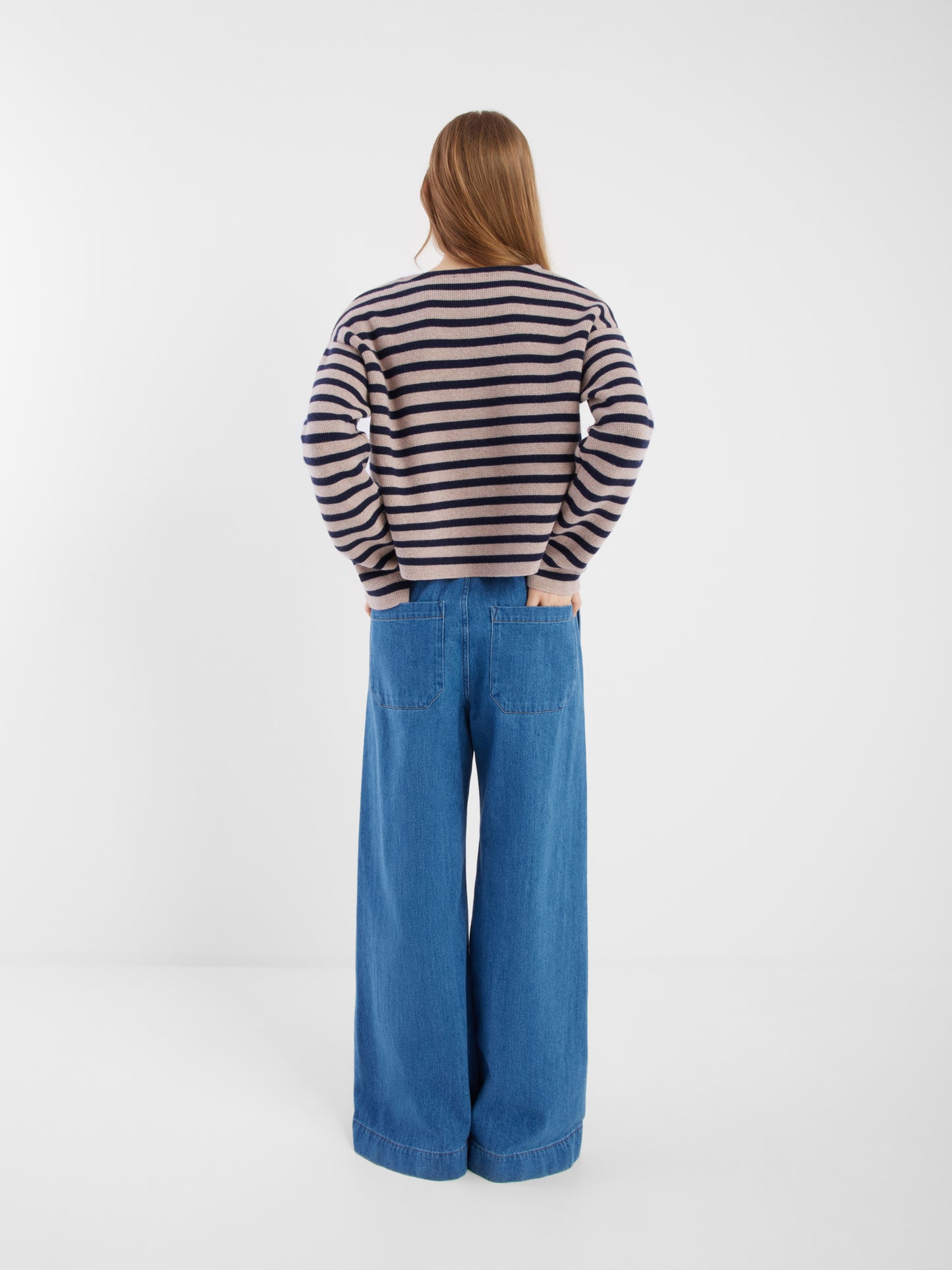 Eine Person mit langen Haaren blickt in die Ferne. Sie trägt die blau-beige gestreifte Jacke Sailor und eine weit geschnittene Bluejeans vor einem schlichten weißen Hintergrund.