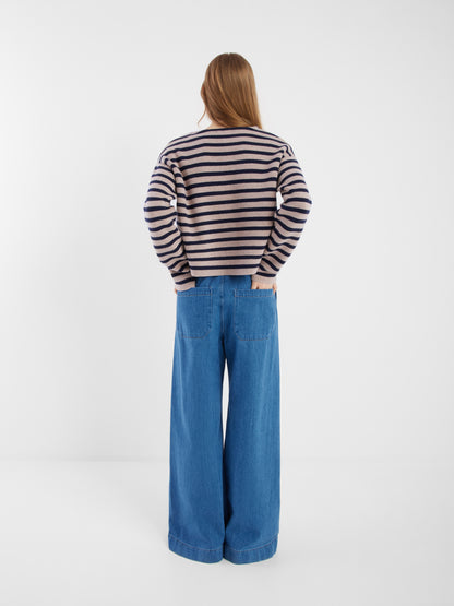 Eine Person mit langen Haaren blickt in die Ferne. Sie trägt die blau-beige gestreifte Jacke Sailor und eine weit geschnittene Bluejeans vor einem schlichten weißen Hintergrund.