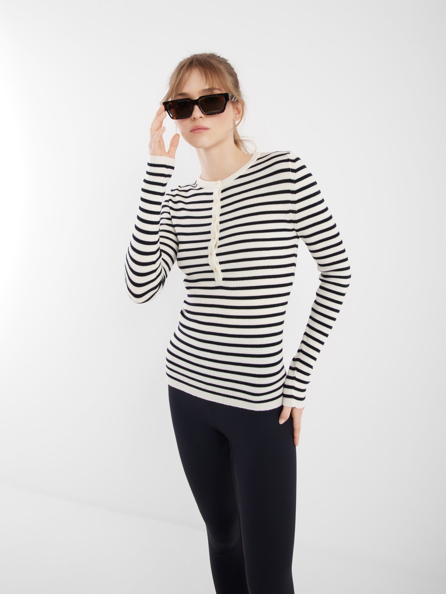 Eine Frau mit schwarzer Sonnenbrille, einem langärmeligen dunkelblauen Strick und schwarzen Leggings steht vor einem schlichten weißen Hintergrund und berührt mit einer Hand ihre Sonnenbrille.