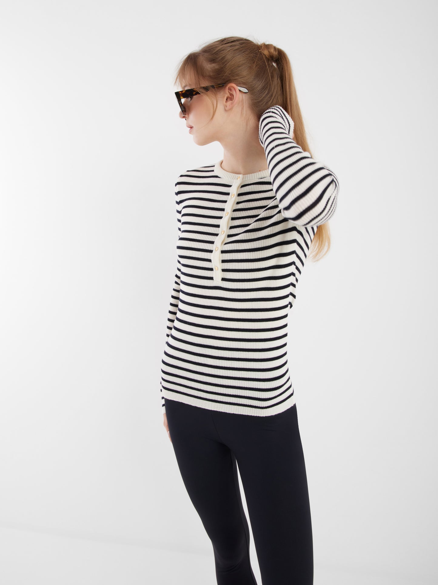 Eine Frau mit langen blonden Haaren in einem Pferdeschwanz trägt eine Sonnenbrille, eine schwarze Hose und den Long Sleeve Strick Dark Blue Stripe. Sie schaut nach links, während sie ihr Haar vor einem schlichten weißen Hintergrund berührt.