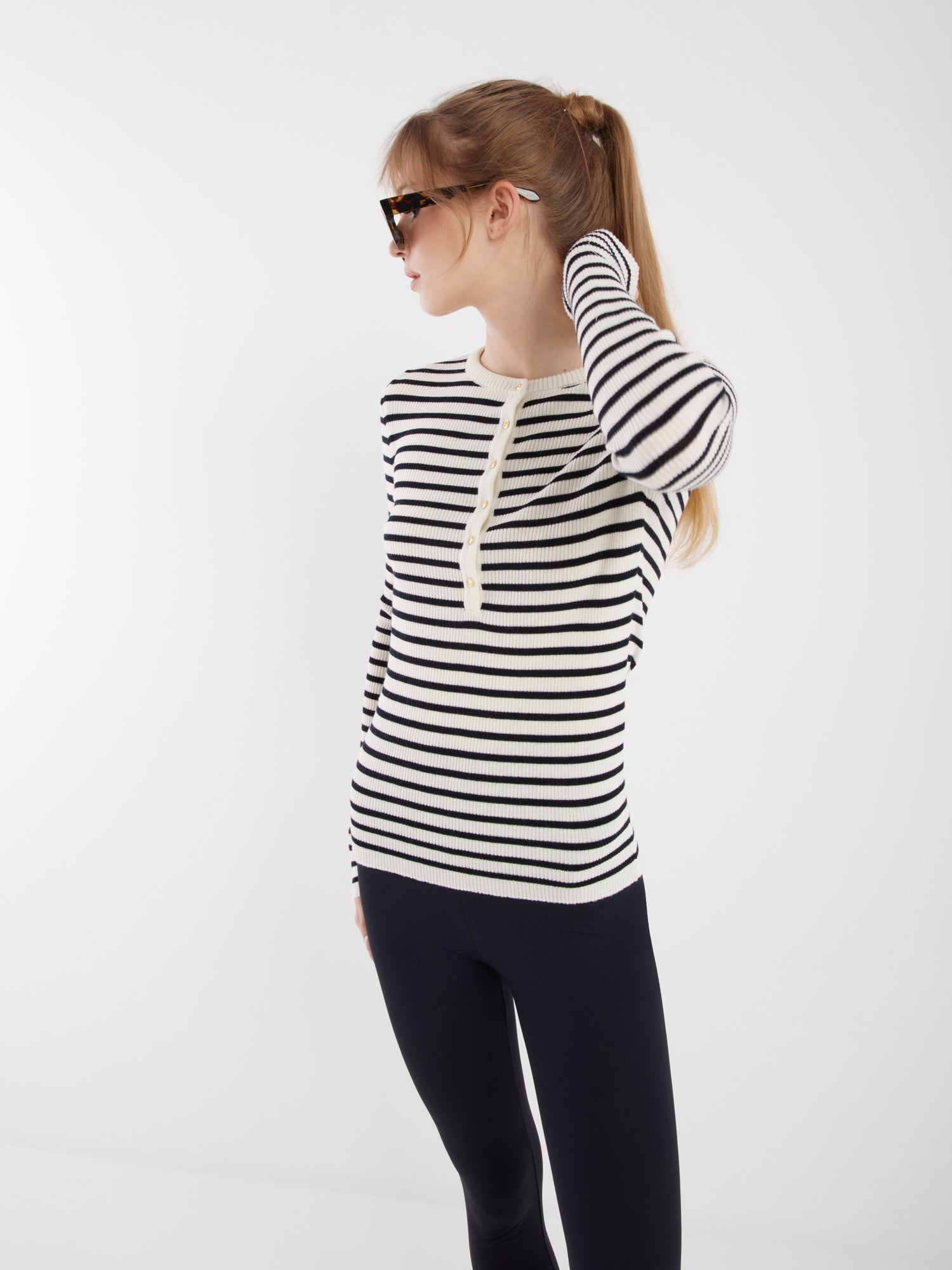 Eine Frau mit langen blonden Haaren in einem Pferdeschwanz trägt eine Sonnenbrille, eine schwarze Hose und den Long Sleeve Strick Dark Blue Stripe. Sie schaut nach links, während sie ihr Haar vor einem schlichten weißen Hintergrund berührt.