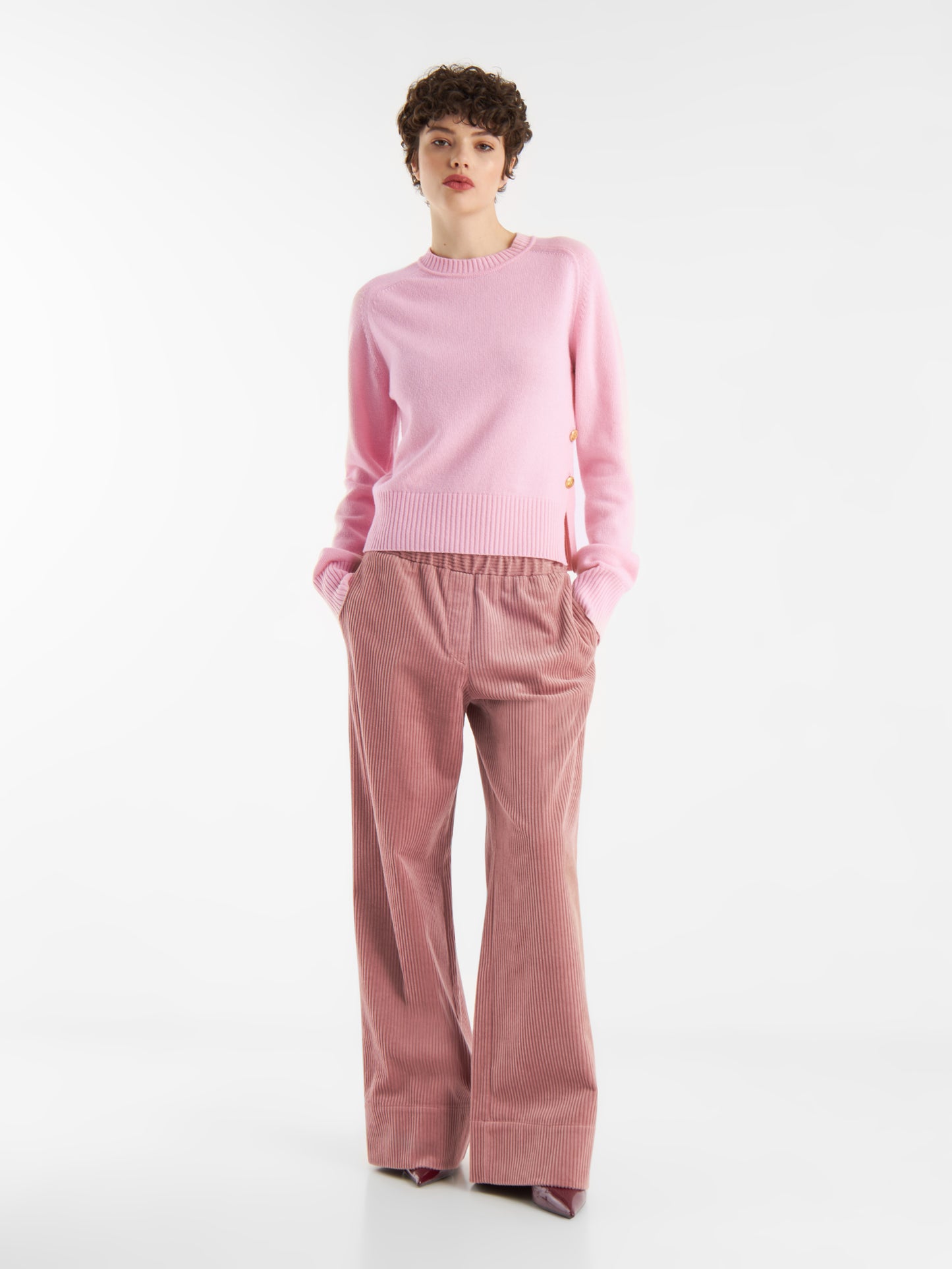 Vor einem schlichten weißen Hintergrund steht eine Person mit kurzen lockigen Haaren, die einen hellrosa Pullover und eine Ted-Rosa-Cord-Hose trägt und die Hände lässig in die Taschen dieser stilvollen Hose steckt.