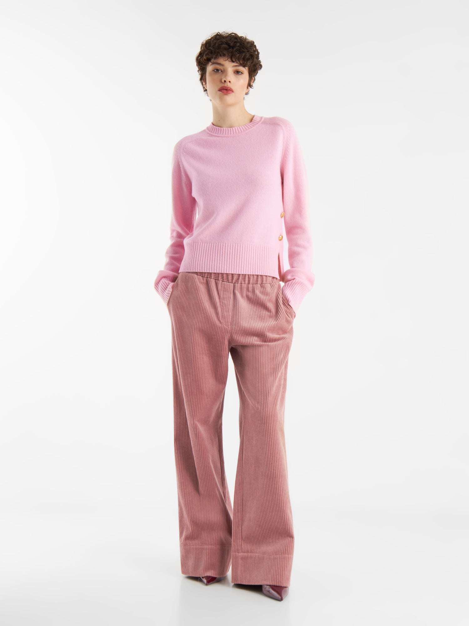 Eine Person steht vor einem schlichten weißen Hintergrund, trägt den Pullover Rosa mit Goldknöpfen und eine lockere, hellbraune Cordhose mit den Händen in den Taschen. Sie hat kurzes, lockiges Haar und blickt leicht nach vorne.