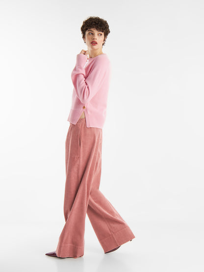 Eine Person, die einen hellrosa Pullover und eine weitbeinige, hochtaillierte Hose Ted Rosa Corduroy trägt, steht seitlich vor einem weißen Hintergrund und blickt mit einer Hand am Kinn über ihre Schulter.