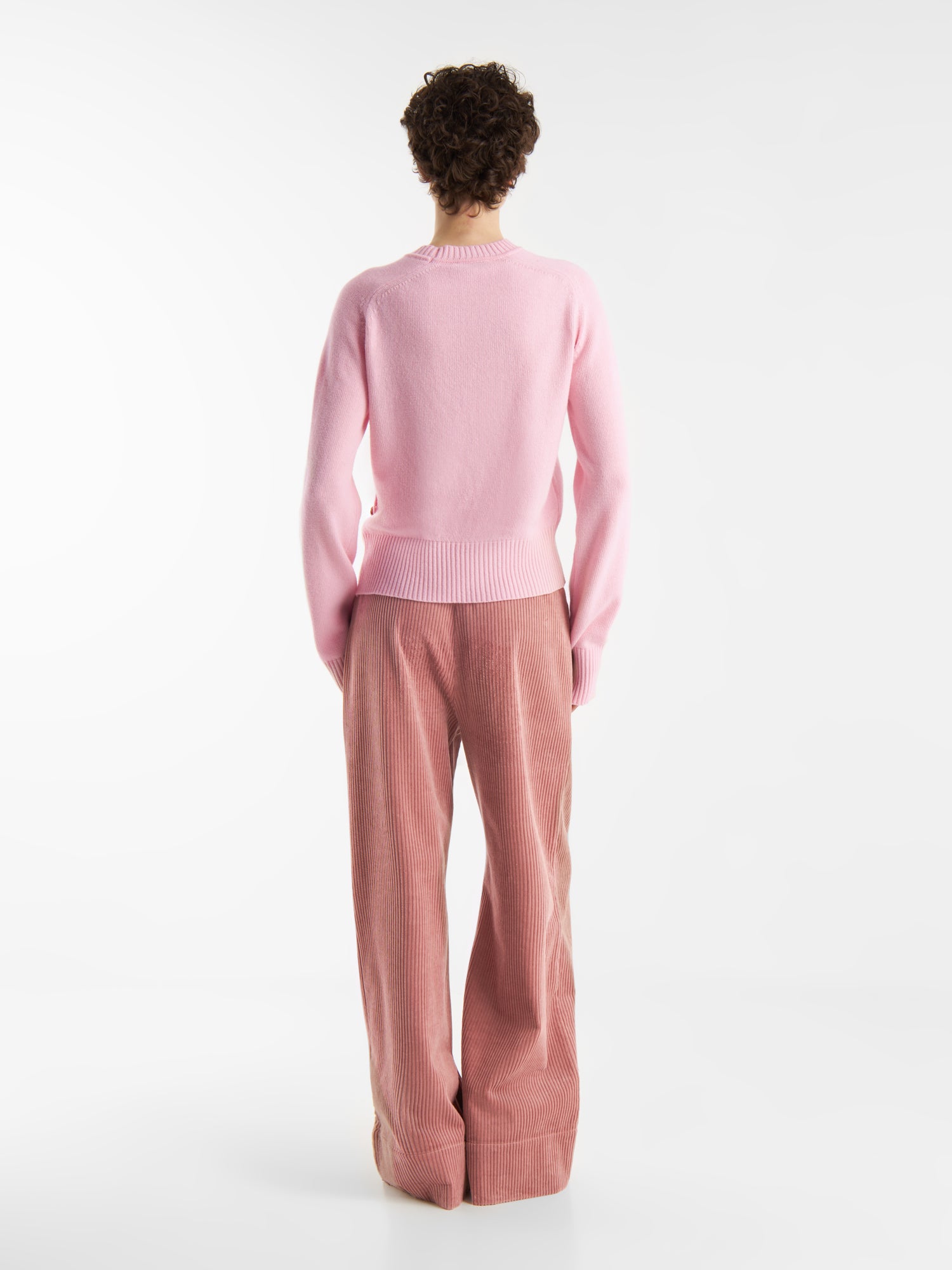 Eine Person mit kurzen, gelockten Haaren steht vor einem weißen Hintergrund und blickt von sich weg. Sie trägt einen hellrosa Pullover und eine locker geschnittene Hose Ted Rosa Corduroy, die den mühelosen Stil der Cordhose Damenmode zeigt.