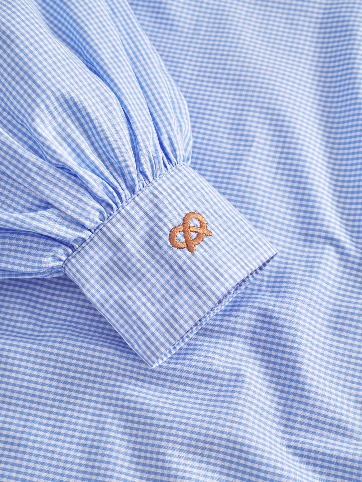 Eine Nahaufnahme der Bluse Antonia Brezel Blue, einem karierten Hemd in limitierter Auflage mit gerafften Ärmeln und einer Manschette, die mit einem braunen Brezelmuster bestickt ist.