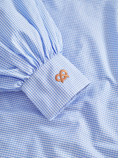 Eine Nahaufnahme der Bluse Antonia Brezel Blue, einem karierten Hemd in limitierter Auflage mit gerafften Ärmeln und einer Manschette, die mit einem braunen Brezelmuster bestickt ist.