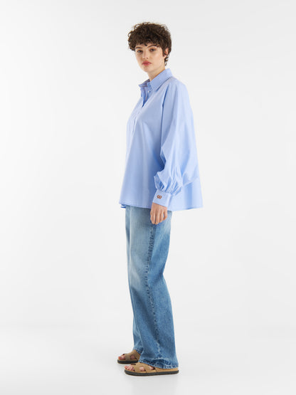 Eine Person mit kurzen lockigen Haaren trägt die Limited Edition der Bluse Antonia Brezel Blue in Übergröße, gepaart mit weit geschnittenen Bluejeans und hellbraunen Sandalen, die seitlich vor einem schlichten weißen Hintergrund stehen.