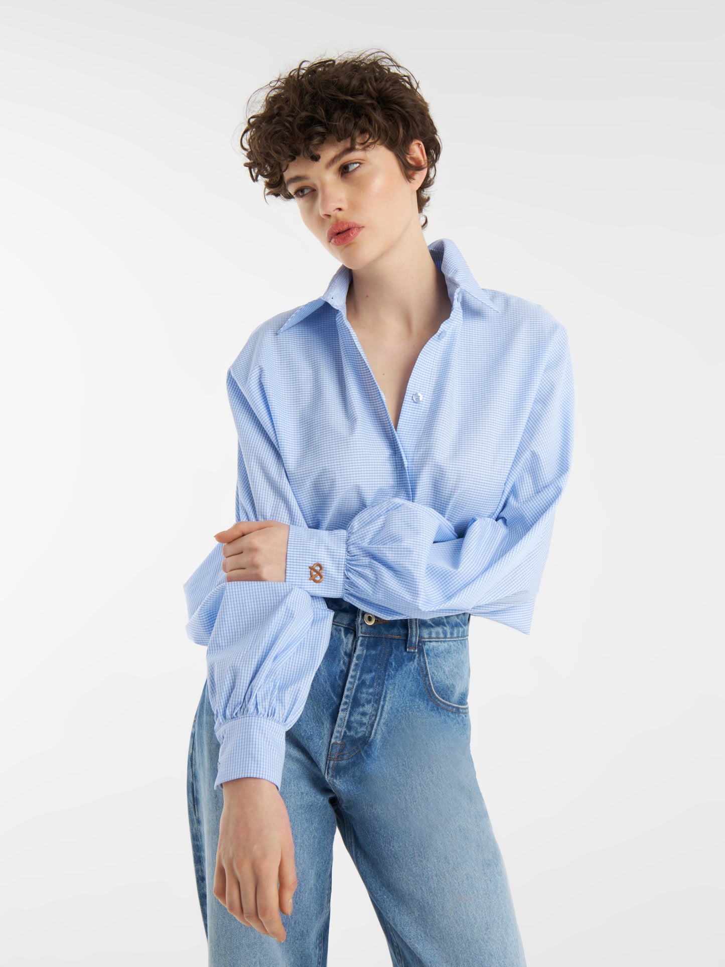 Eine Person mit kurzen lockigen Haaren trägt die Bluse Antonia Brezel Blue, eine limitierte Auflage der Bluse Antonia Brezel Blue in Hellblau mit hochgekrempelten Ärmeln, gepaart mit einer hochgekrempelten Blue Jeans, vor einem schlichten weißen Hintergrund.