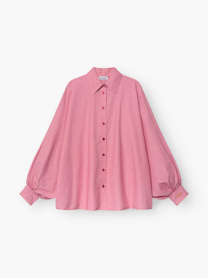 Die Bluse Antonia Brezel Red ist eine locker sitzende, langärmelige rosafarbene Bluse mit Halsausschnitt, Knopfleiste, Puffärmeln mit geknöpften Manschetten und einem kleinen gelben Etikett an einer Manschette.