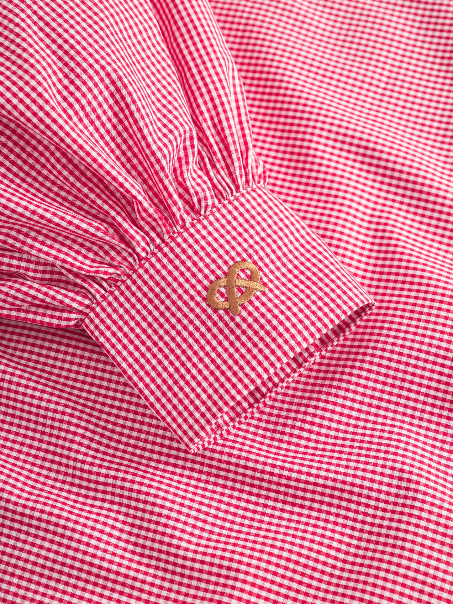 Eine Nahaufnahme des roten Ärmels der Bluse Antonia Brezel, die die rot-weiß karierte Manschette mit goldener Symbolstickerei hervorhebt.