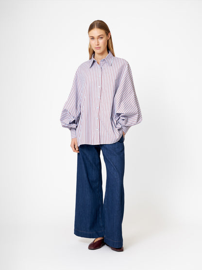 Eine Frau mit glattem, langem Haar und neutralem Gesichtsausdruck steht vor einem weißen Hintergrund und trägt die Bluse Antonia Blue Burgundy Stripe mit weiten Ärmeln, weite Bluejeans und braune Schuhe.