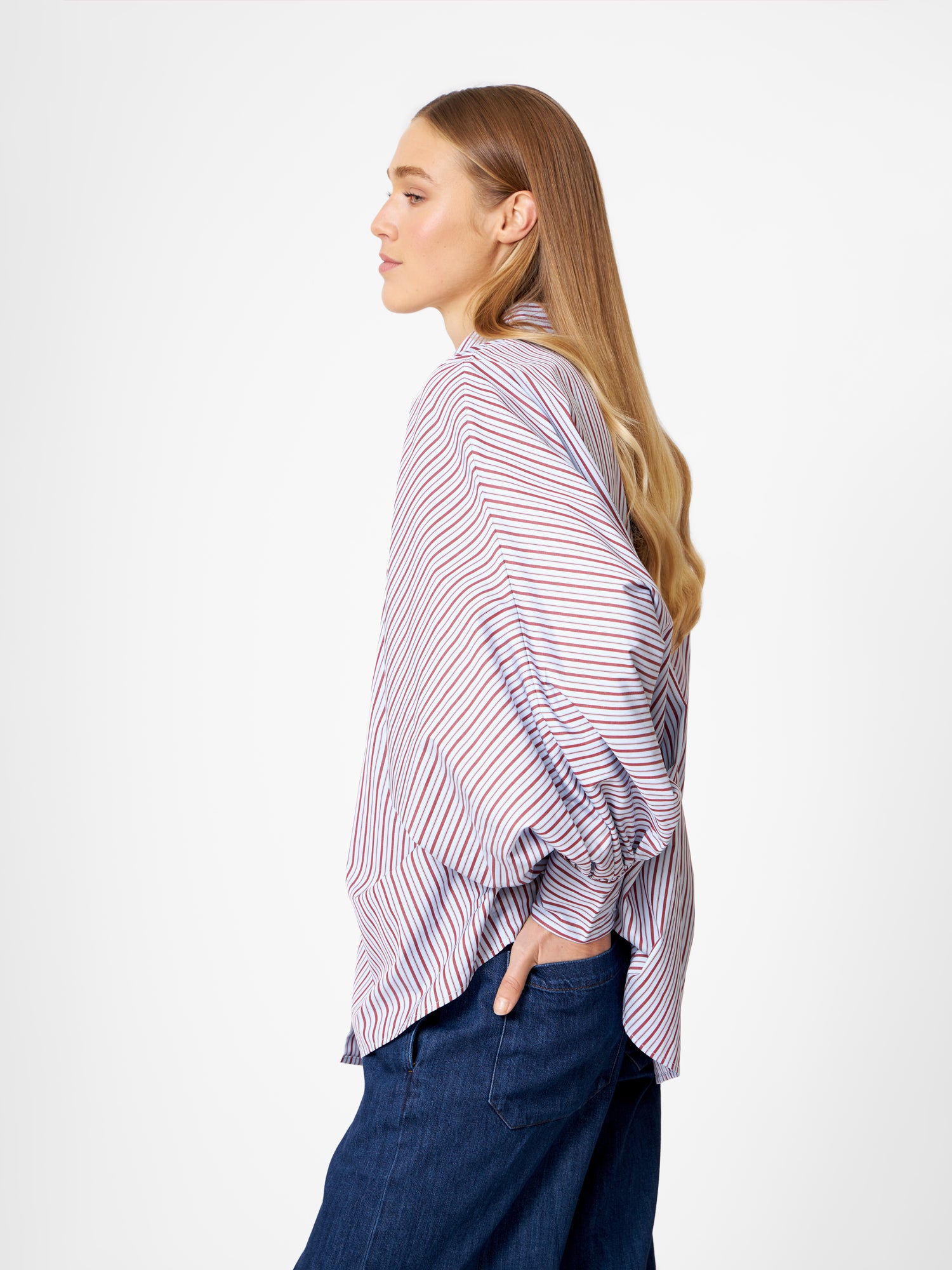 Eine Frau mit langem, glattem blondem Haar steht im Profil vor einem weißen Hintergrund. Sie trägt die Bluse Antonia Blue Burgundy Stripe und blaue Jeans und hat eine Hand in der Tasche.